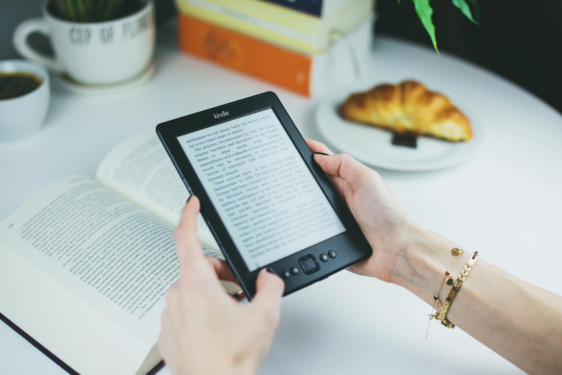 Kobieta czyta na Kindle przy stole z książką i rogalikiem